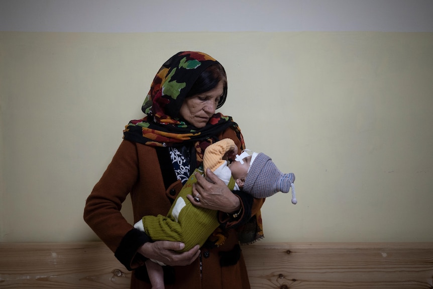 A Middle Eastern grandmother with headwear looks at a baby that she holds in hospital ward