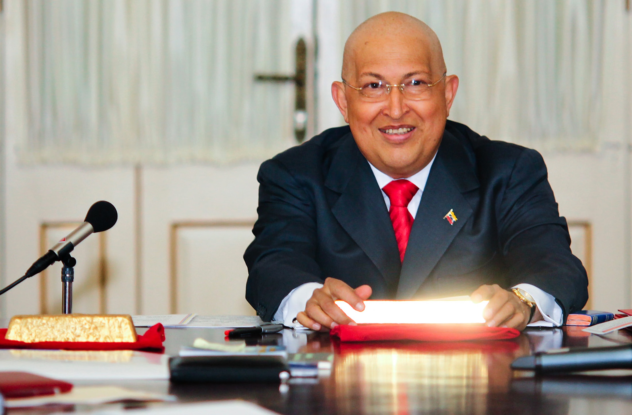 A bald Hugo Chavez wearing a dark suit and red tie, sitting while smiling and holding a shining gold bar ingot on a red cloth.