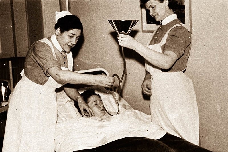A black and white image of two nurses holding a funnel and tube to a patient lying down with their eyes closed.