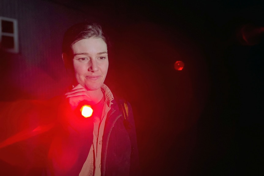 A woman with brown hair holds a red torch light in the dark 