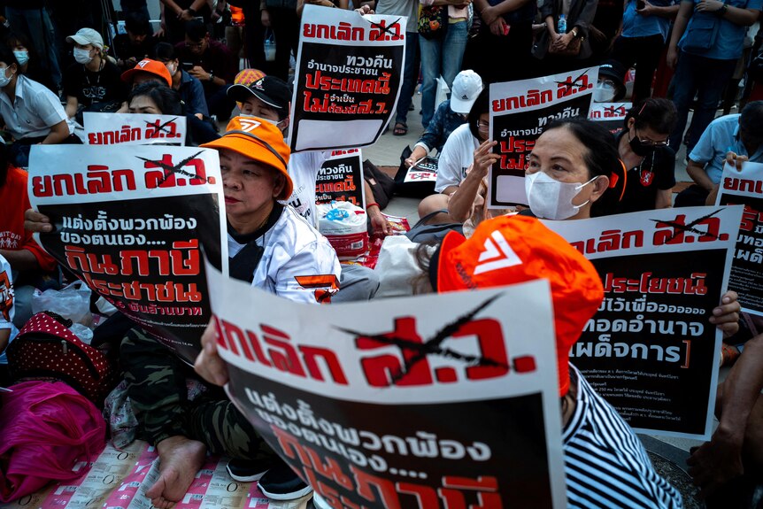 A group of protesters sit on the ground, holding signs with writing in Thai
