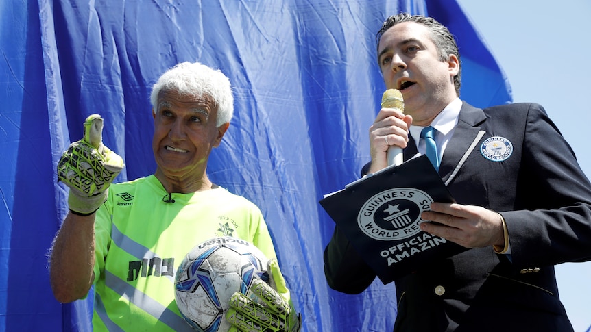 Isaak Hayik points off camera whilst holding a soccer ball, standing next to a man in a suit with a microphone and clipboard