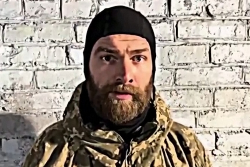Бородатый мужчина в черной шляпе стоит перед белой кирпичной стеной.