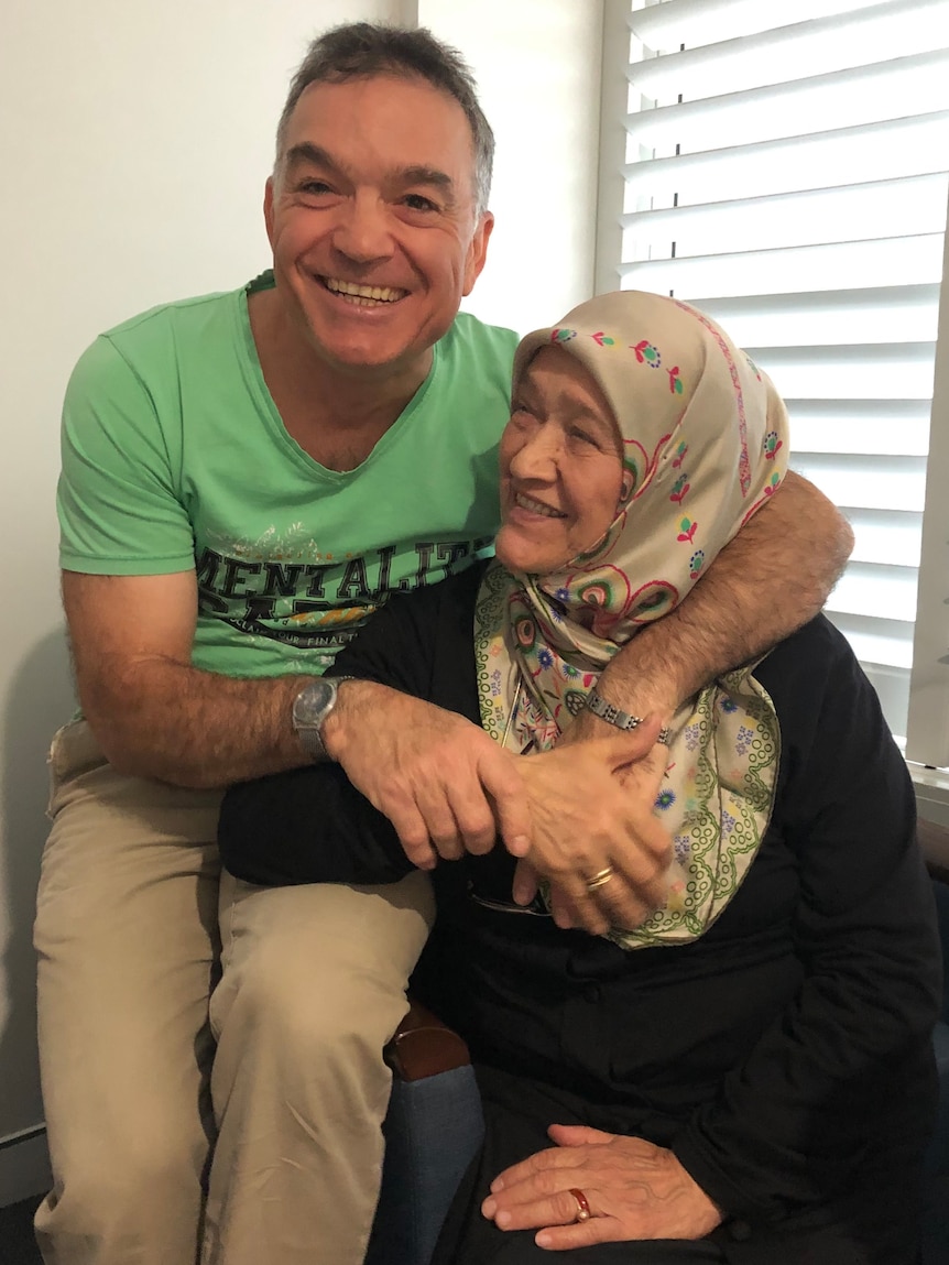 A man in a green t-shirt hugs his mother who looks at him smiling. 