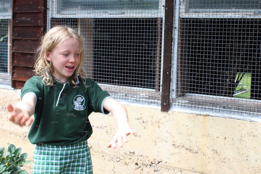 A blond primary school girl in a green uniform, standing outside with her arms out.
