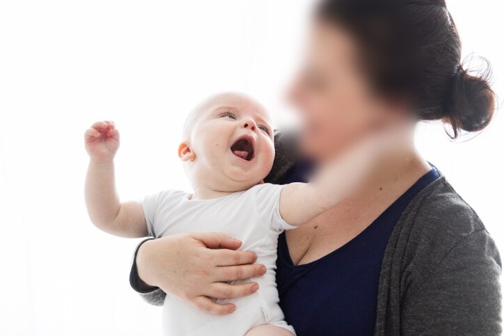 A woman whose face is blurred holds a baby. The baby is looking up at her face with his mouth open and arms out.