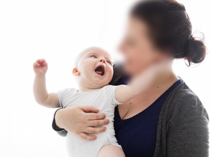 A woman whose face is blurred holds a baby. The baby is looking up at her face with his mouth open and arms out.