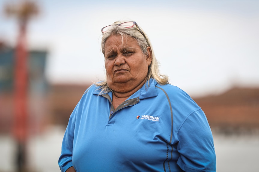 An older, large, Indigenous woman stares with a stern facial expression , glasses on top of her head, turquoise jumper.