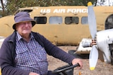 A man in a jacket and plait shirt looks off to side while a propeller and plane marked US Air Force sits in background 