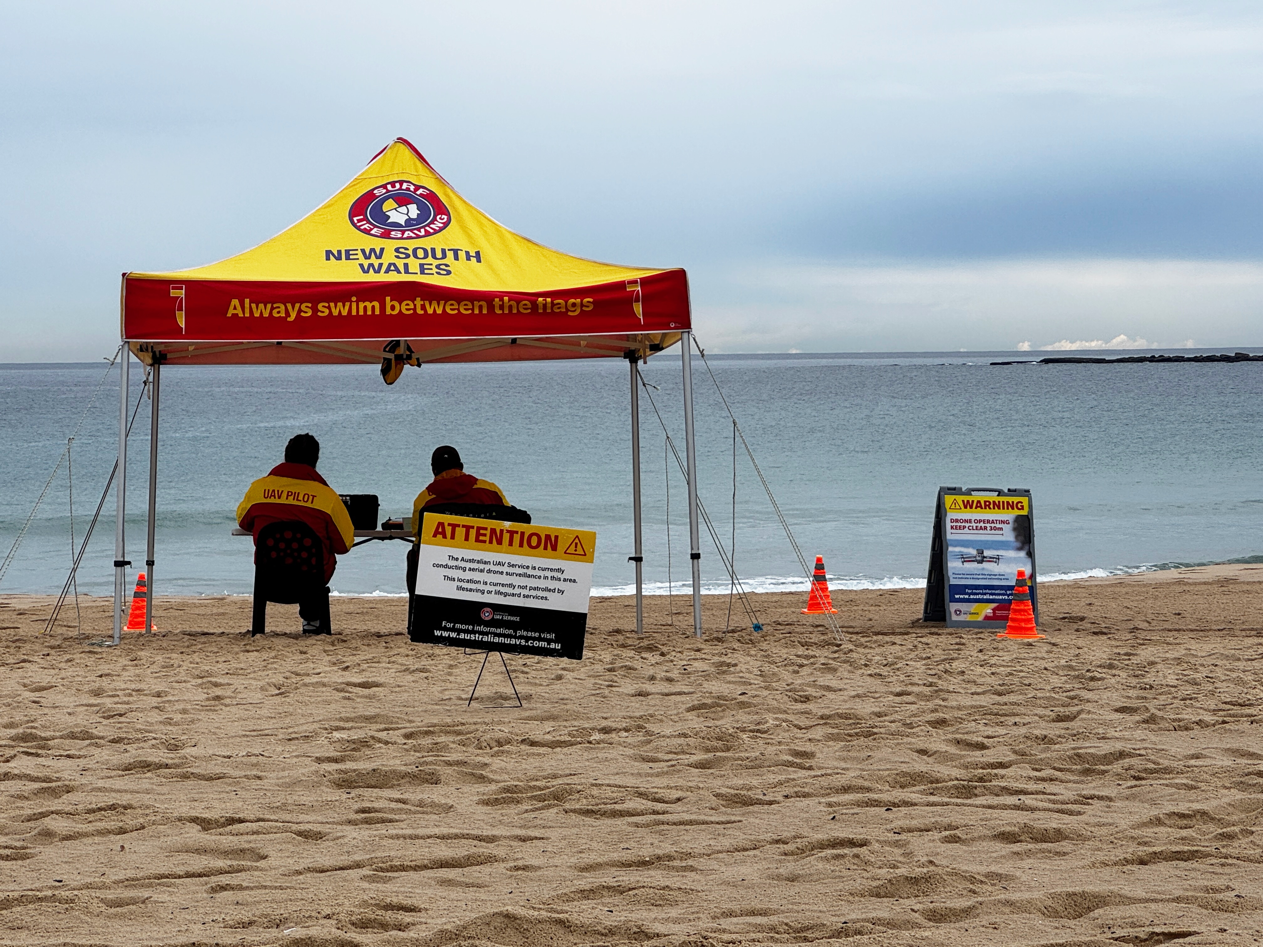 A surf life saving tent with two people in uniform under it with signs saying drone surveillance is happening.