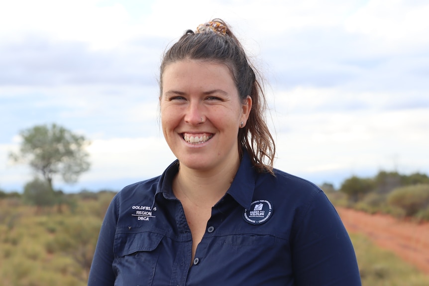 A smiling young woman in dark coveralls stands in the desert.