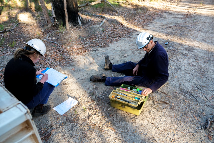 A man sitting on the forest floor near a tackle box full of items. A woman is crouched nearby taking notes.