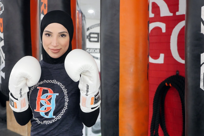 A female boxer wearing a hijab looks at the camera, posing with her boxing gloves in front of her, and punching bags behind.