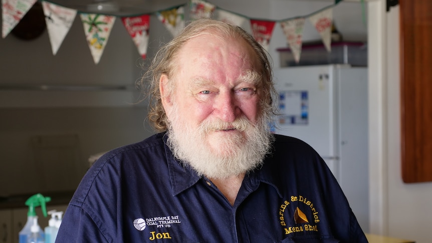 Jon Eaton, an elderly man stands infront of the Men's Shed Kitchen smiling and wearing a blue workers shirt with his name on it.