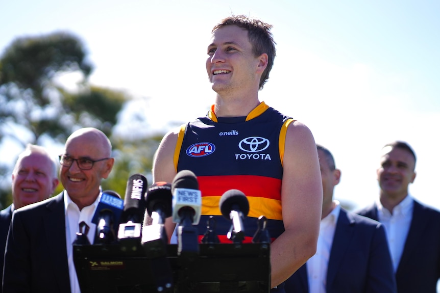 A smiling man wearing a Crows guernsey stands in front of media microphones. Behind him are a group of smiling men in suits