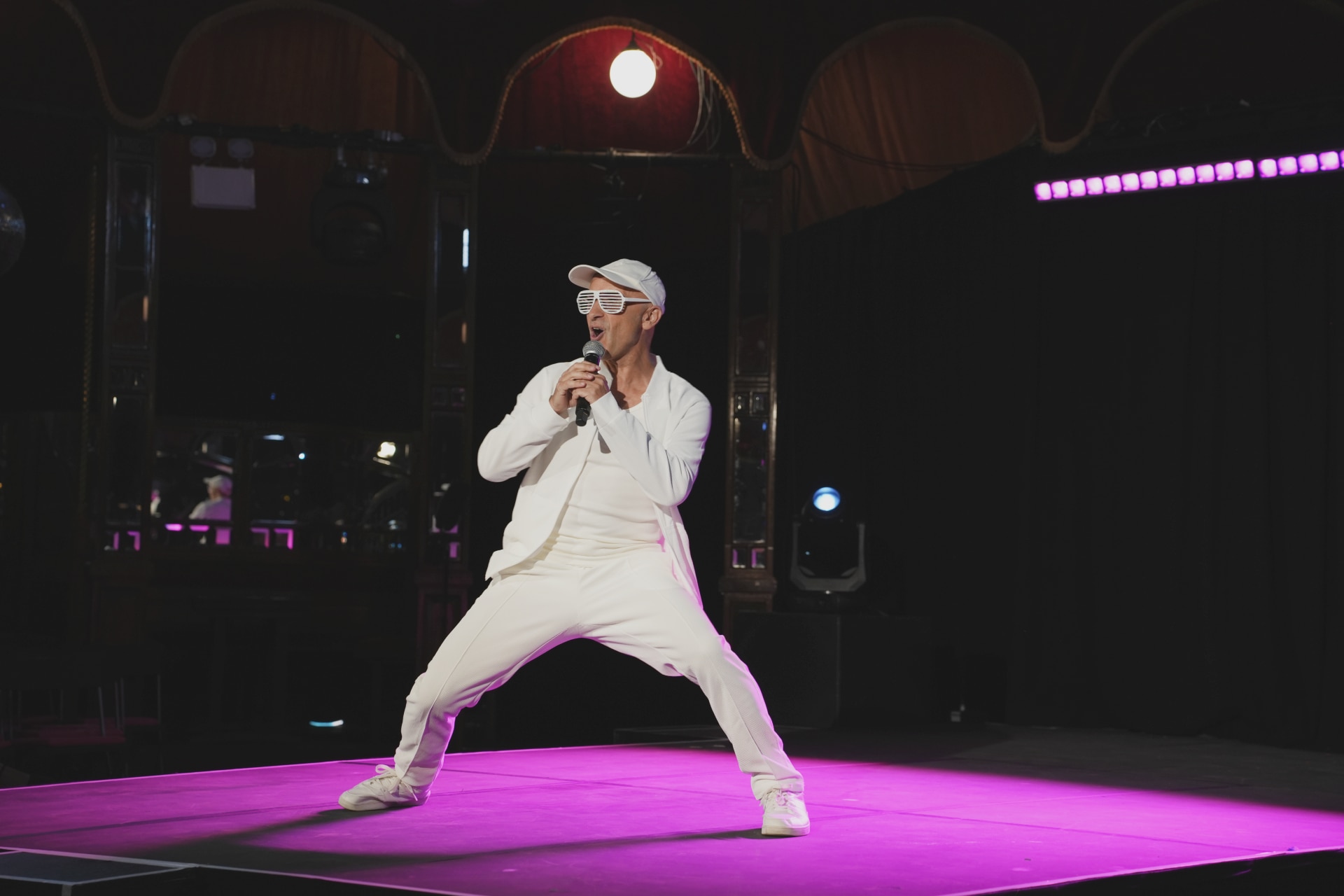 Un hombre con traje blanco, gorra y gafas posa durante un espectáculo. 