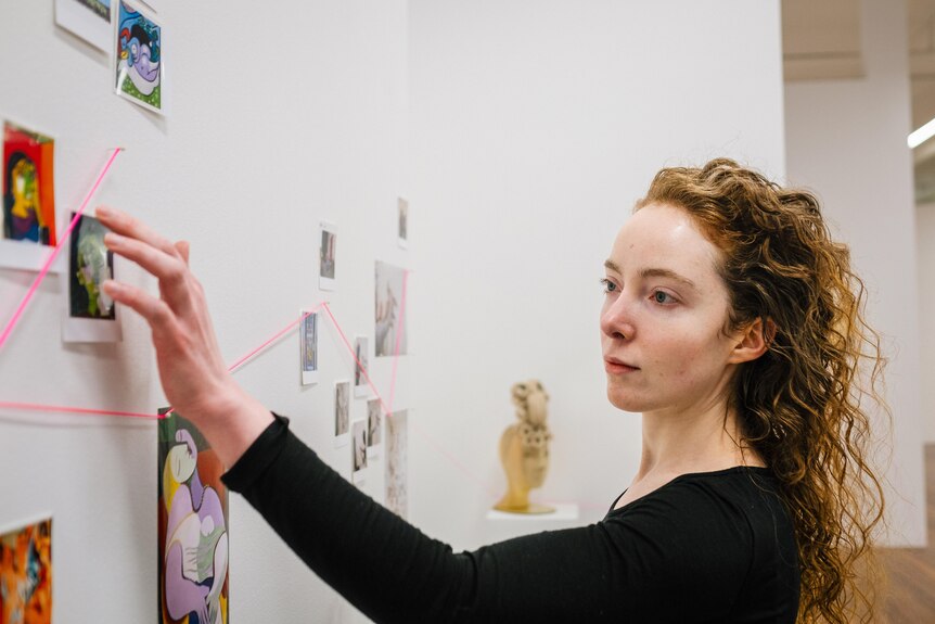 Picture of artists face gazing and reaching out at the wall in her studio. She has long red curly hair and wears a black shirt