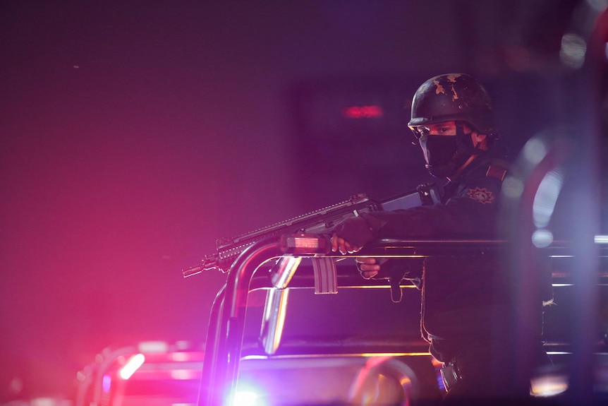 A police officer with helmet, mask and gun stands on vehicle and looks down at night with light behind him