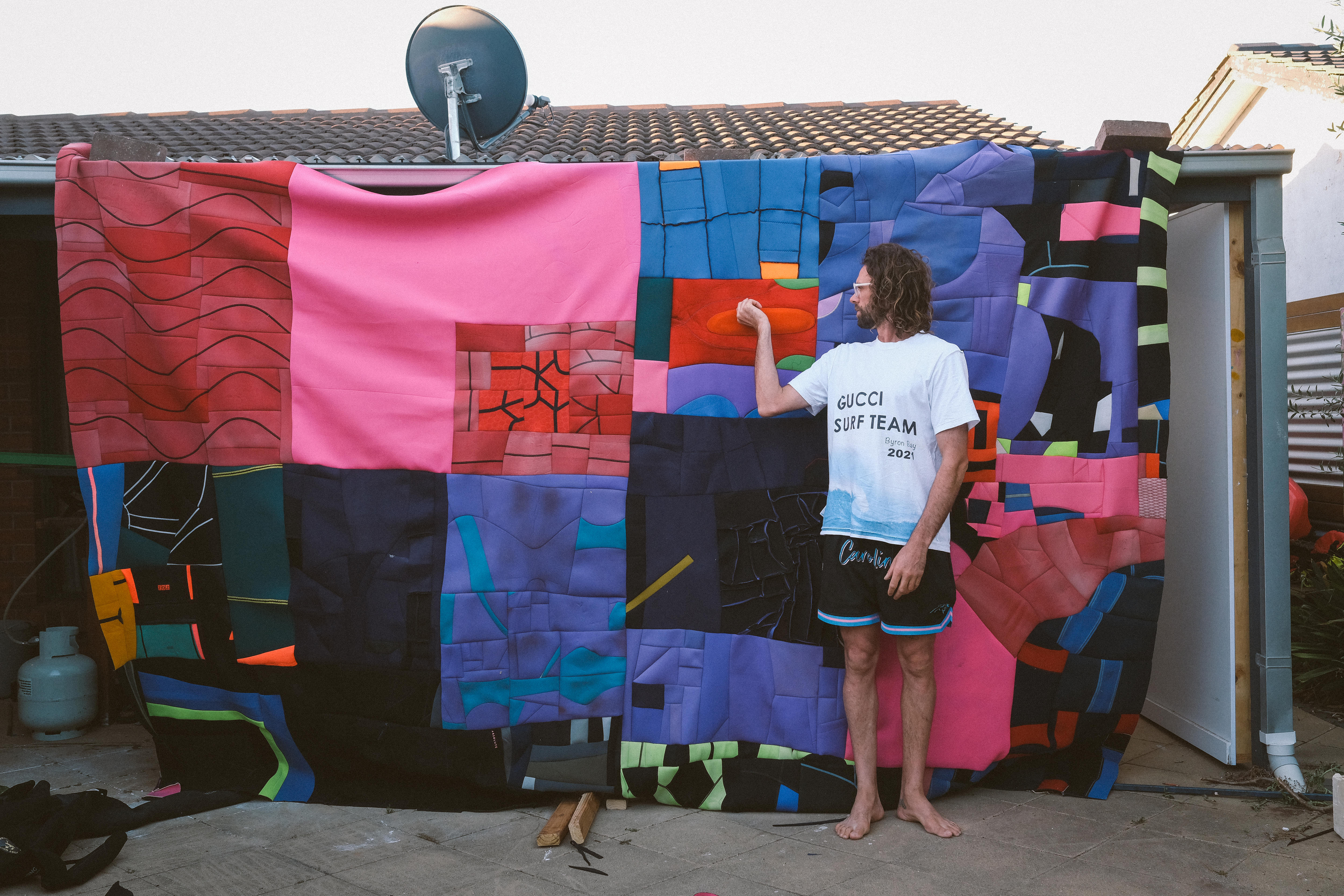 Artist stands in backyard in front of a large painting hanging up, made out of wetsuit fabric