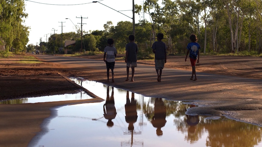 Four children walk down a street in Doomadgee, they are reflected in the water of a flooded gutter.