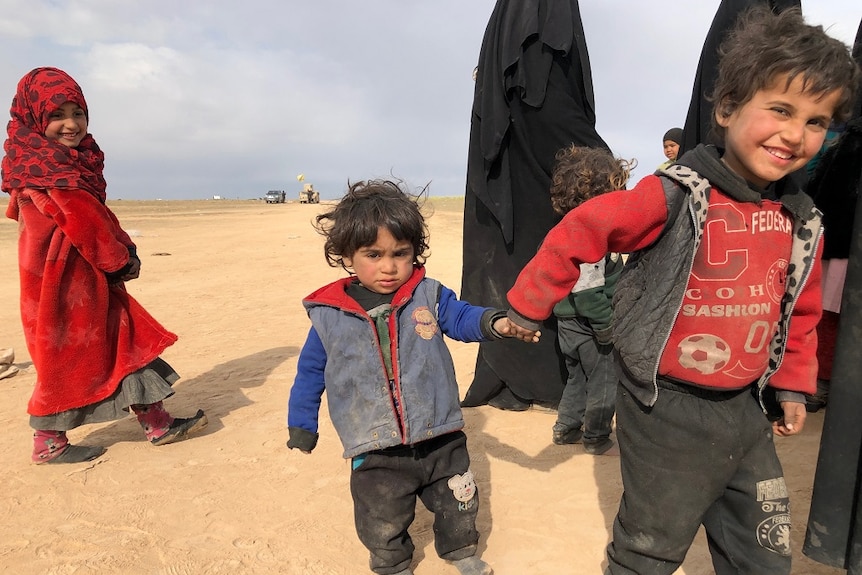 A young boy smiles as he holds the hand of a younger boy and leads him as they walk across an open area with a group