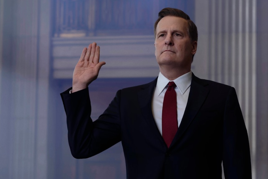 Jeff Daniels as James Comey, in a suit and tie, looks serious as he holds his right palm in the air while taking an oath
