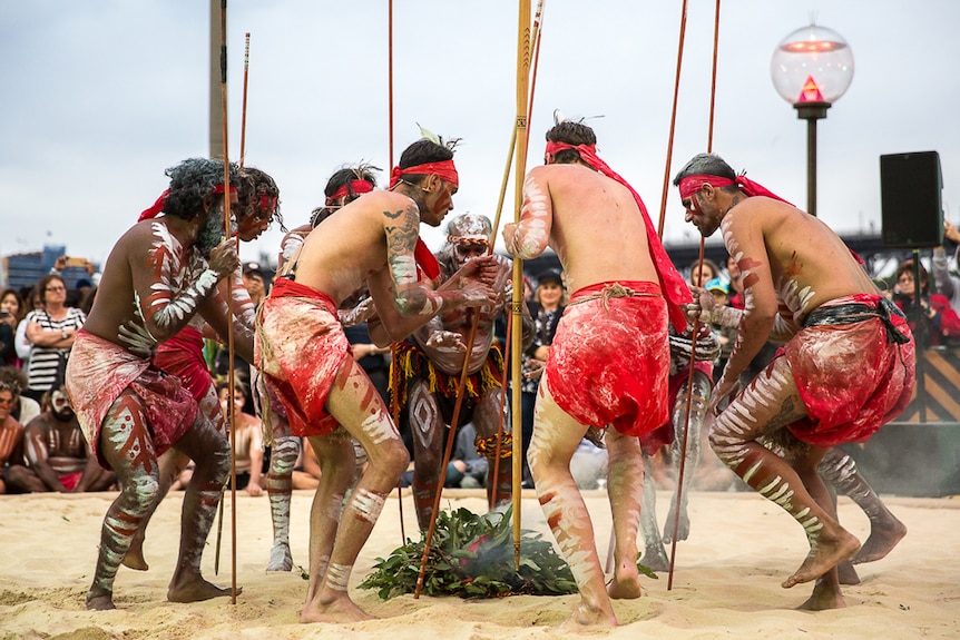 Aboriginal dancers holding spears huddled around a fire covered with gum leaves.