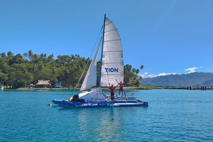 A catamaran in a tropical harbour. Its crew stand with their arms raised in excitement.