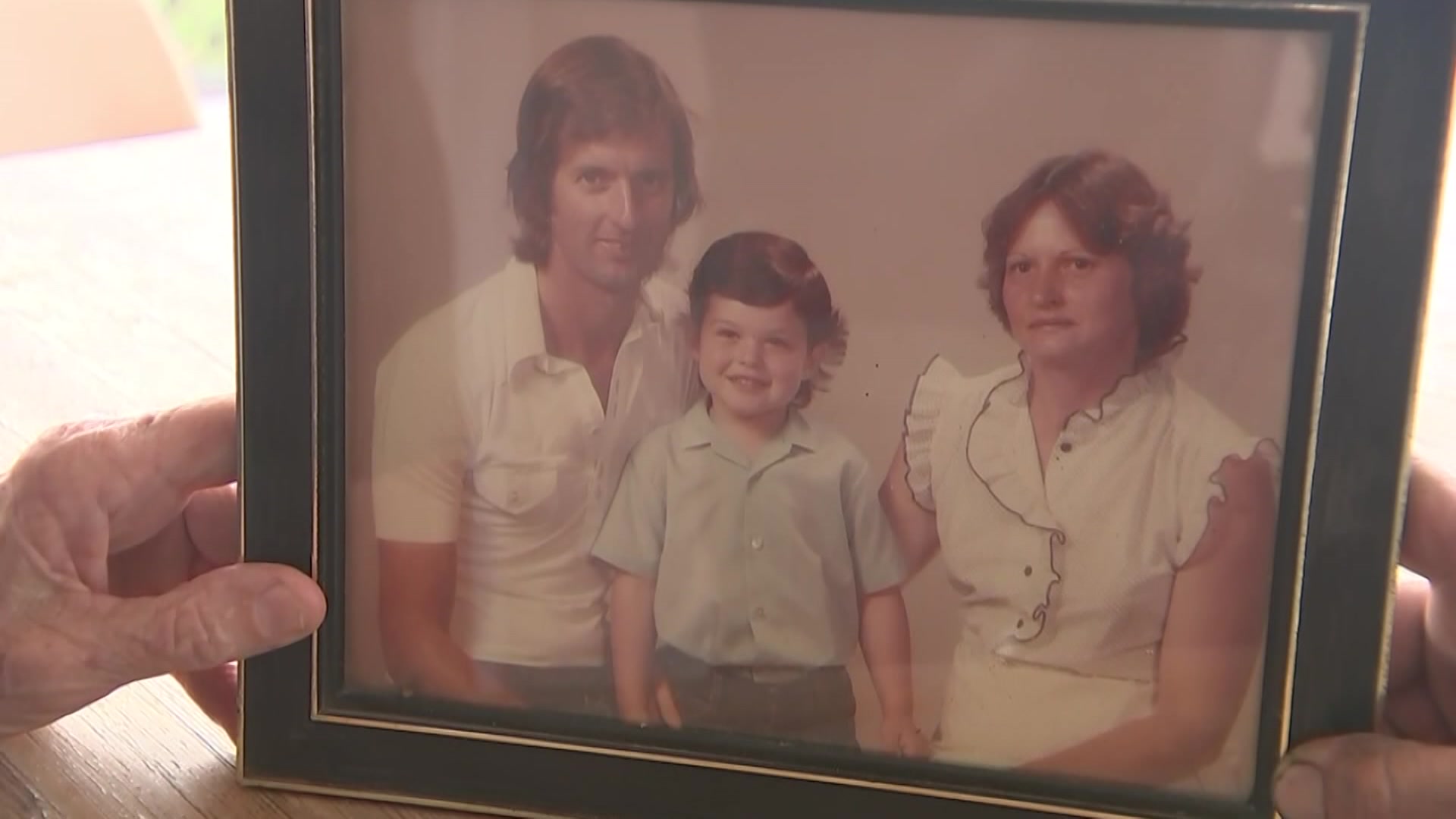 Hands hold an old, sepia-toned photo of a child with parents.