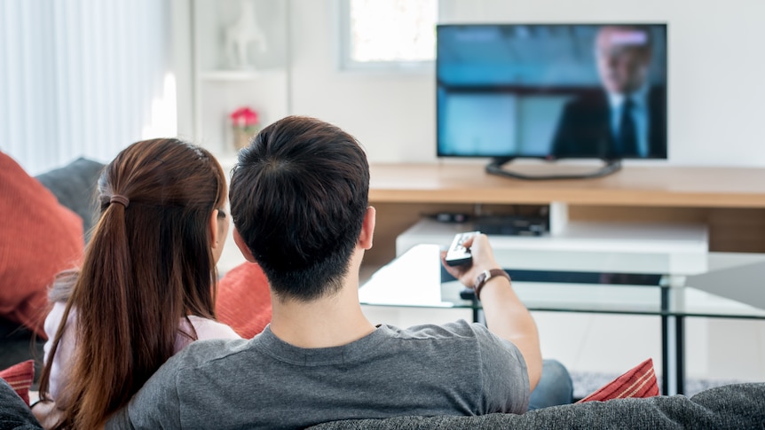 A couple sit watching TV, the man holds up a remote control