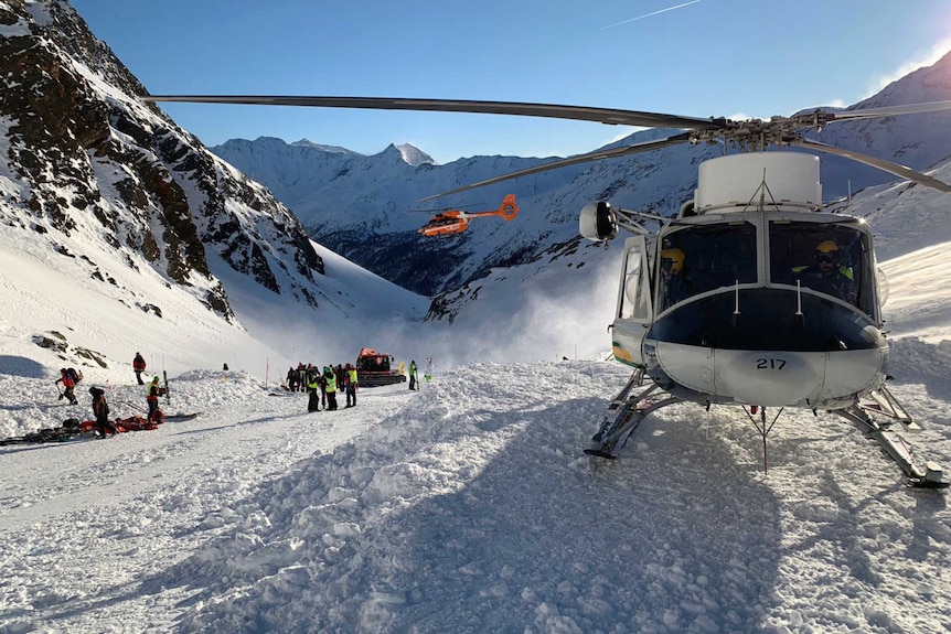 A helicopter sits on a snowy mountain while a group of people in bright red and yellow ski gear congregate behind.