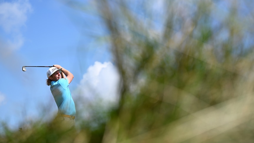 A man plays a golf shot from behind long grass.