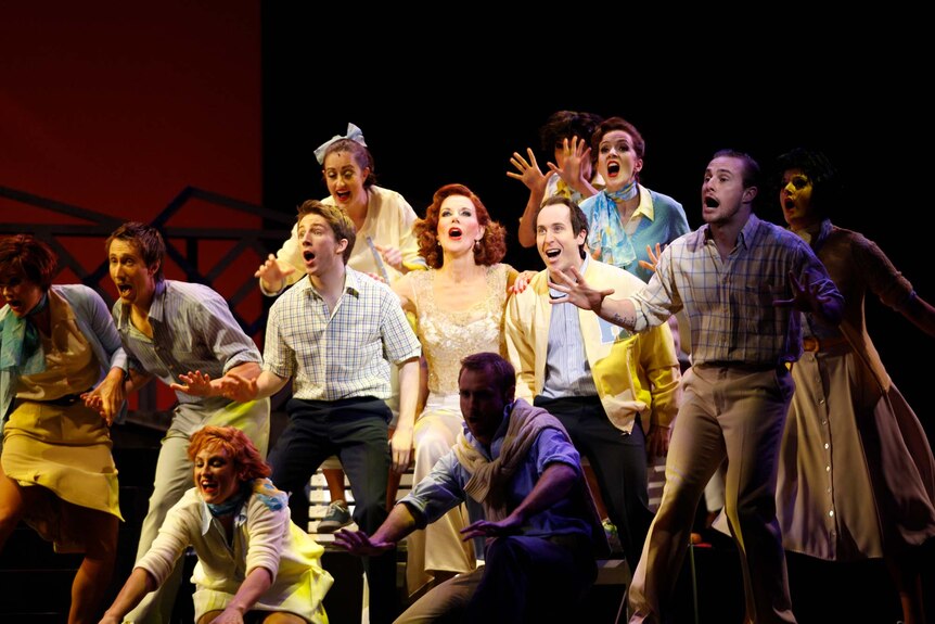 On stage, a cast of 12 performers in 1960s style daywear sing enthusiastically.