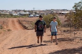Two children walking along a red dirt track with buildings in the background