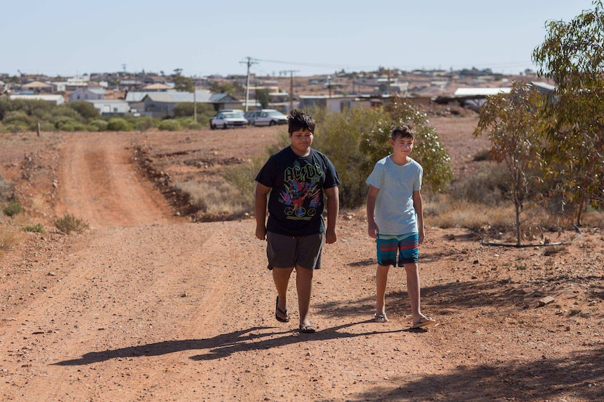 Two children walking along a red dirt track with buildings in the background