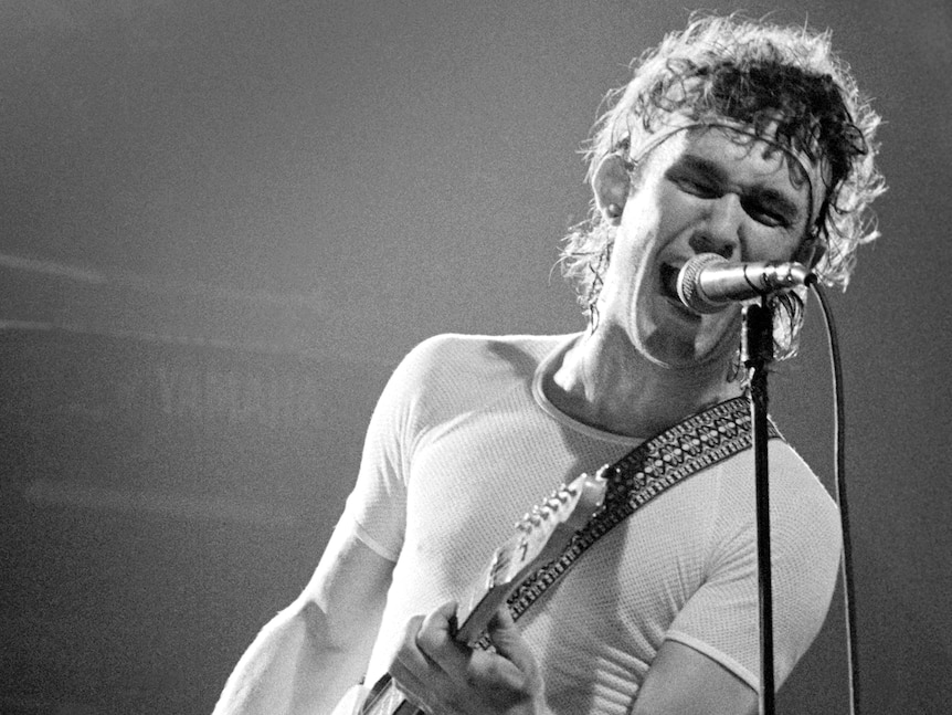 Black and white photo of young Jimmy Barnes with headband and tight, white shirt yelling into microphone and holding guitar.