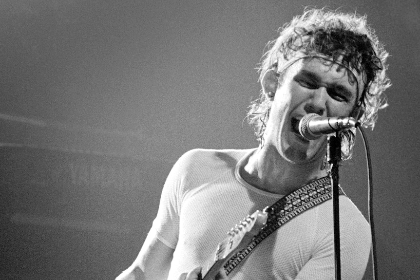 Black and white photo of young Jimmy Barnes with headband and tight, white shirt yelling into microphone and holding guitar.