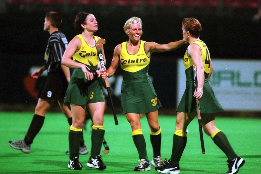 Three players from the Australian Hockeyroos, wearing green and gold uniforms, celebrate a goal.