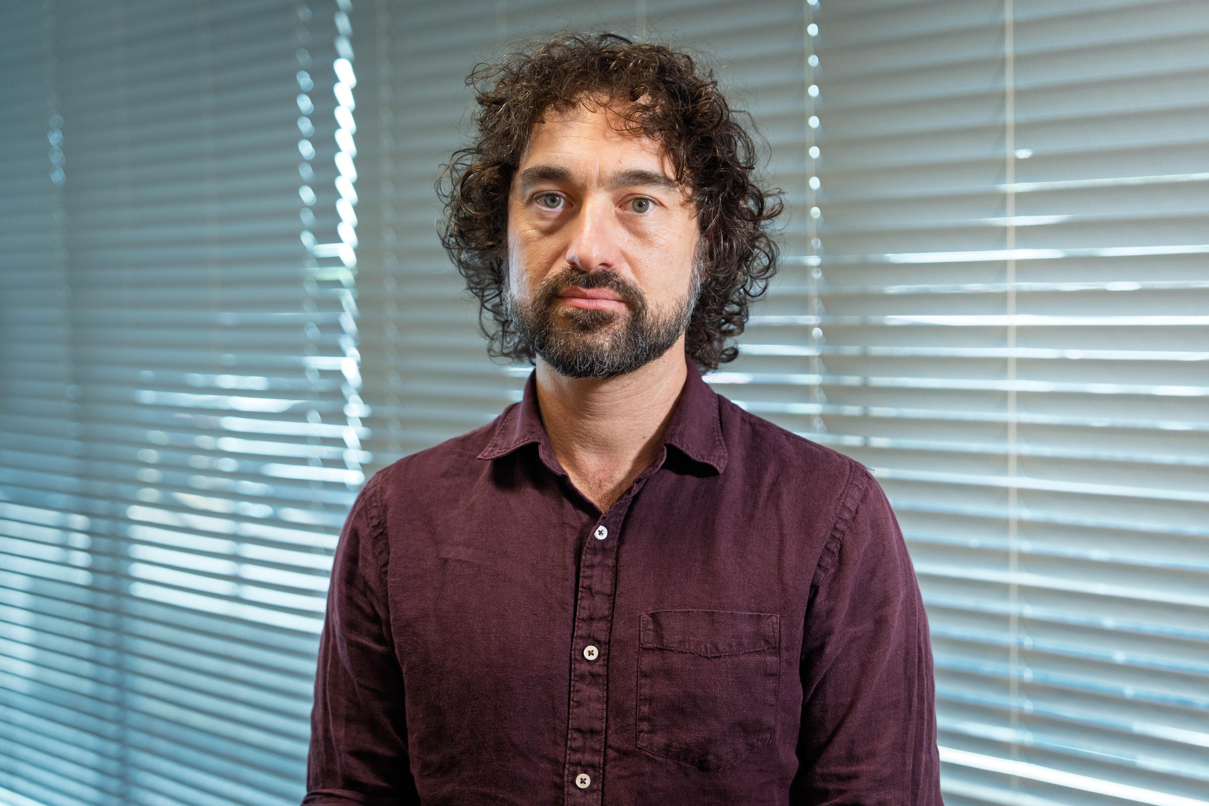 A man with black hair and a purple shirt looks at the camera 