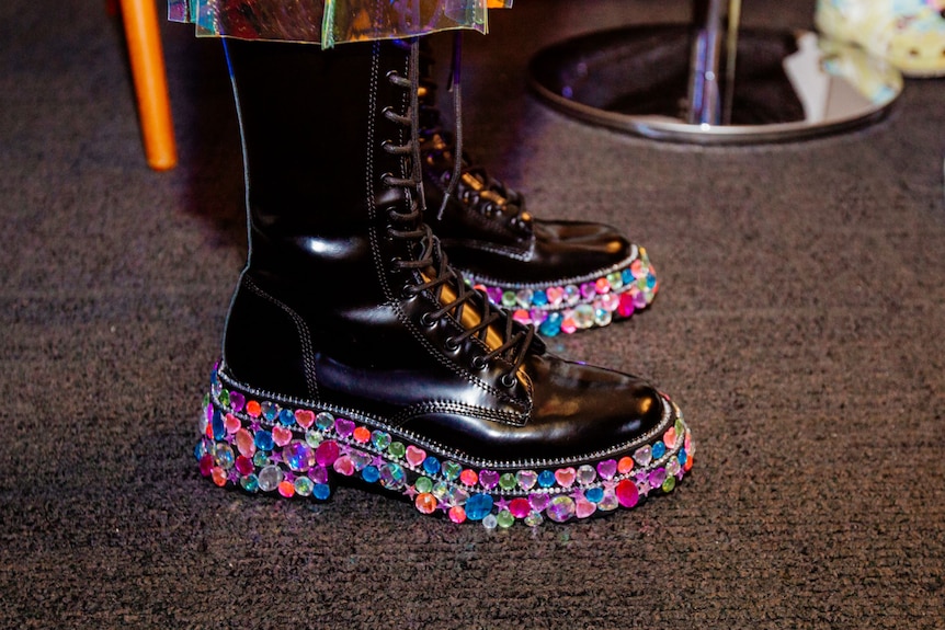 Black boots with feet in them are seen in a closeup shot. The shoes are bedazzled with multicolour rhinestones.