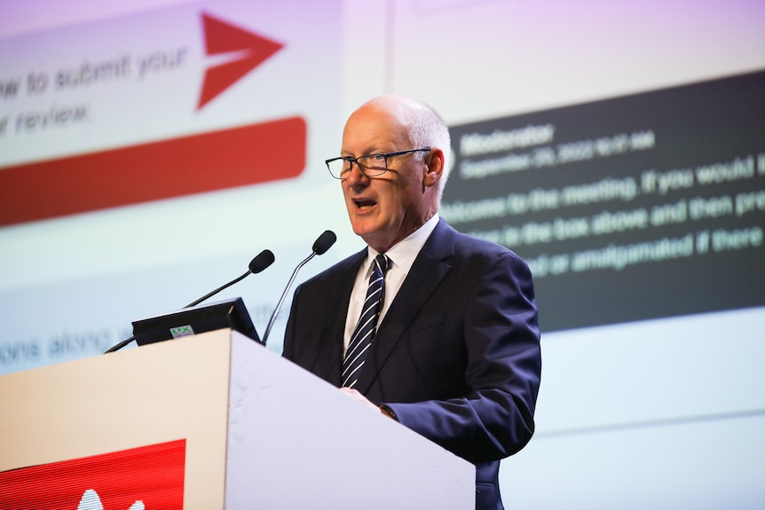 A man with glasses and a navy blue suit and tie stands behind a lecturn at a business conference