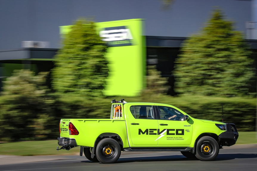 A green ute drives in front of a blurry green garden