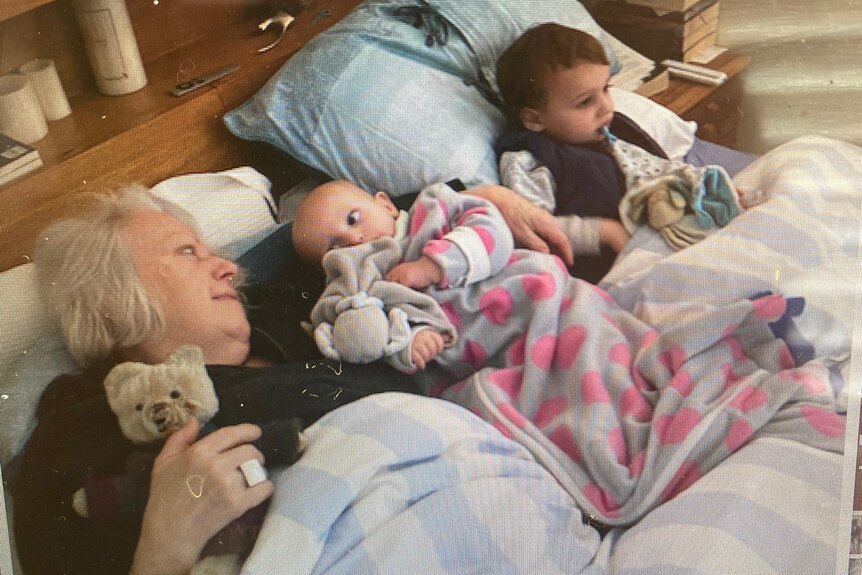 An older woman lays in bed with two small children, all cuddling their teddies.