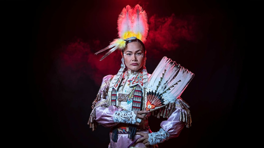 A woman wears pale pink Native American regalia decorated with cone bells holds intricately decorated feather fan up to shoulder