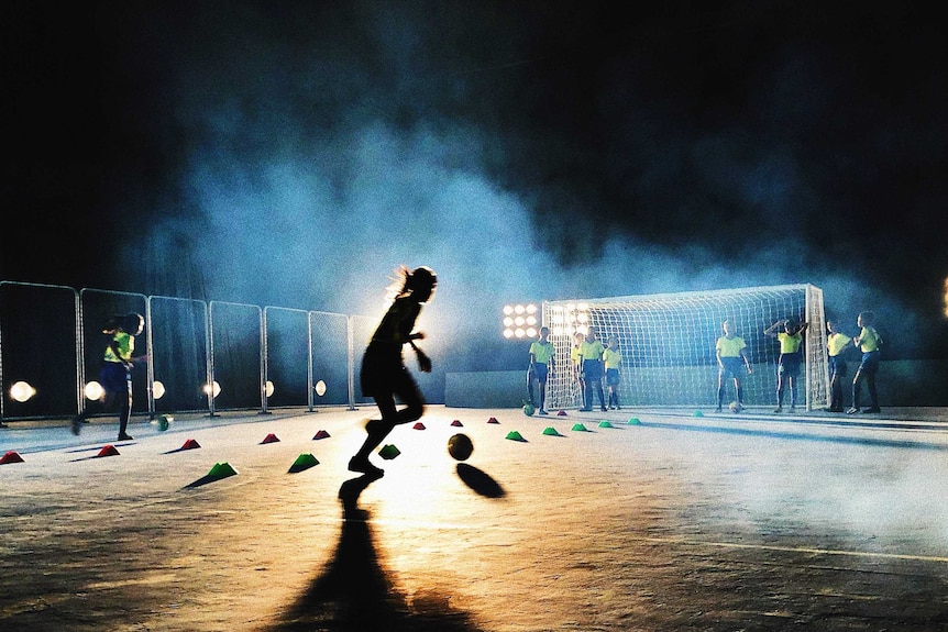 A team of people takes turns to practice soccer at the net, at night time.