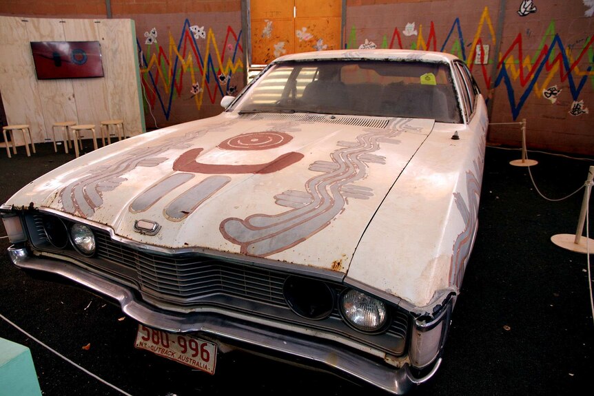 A white 1973 Ford Fairlane with brown and blue water dreaming paintings on it.