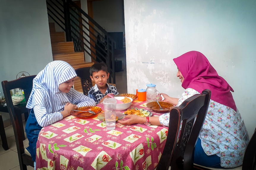 A girl in a veil sits at a dinner table with a small boy and a woman in a veil