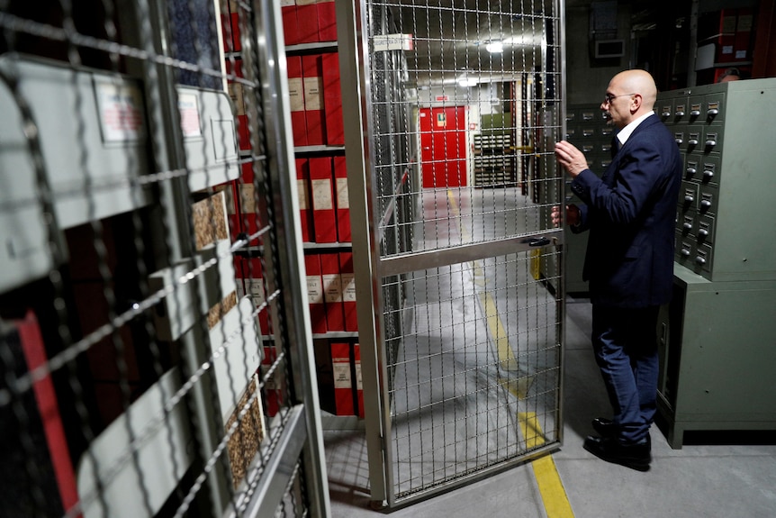 Man stands by wire gates leading down a hall that ends in a red door 