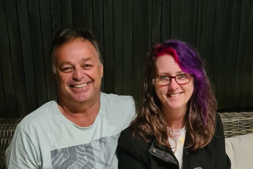 A man and a woman, he in a blue tshirt smiling and her with a purple section of hair with glasses