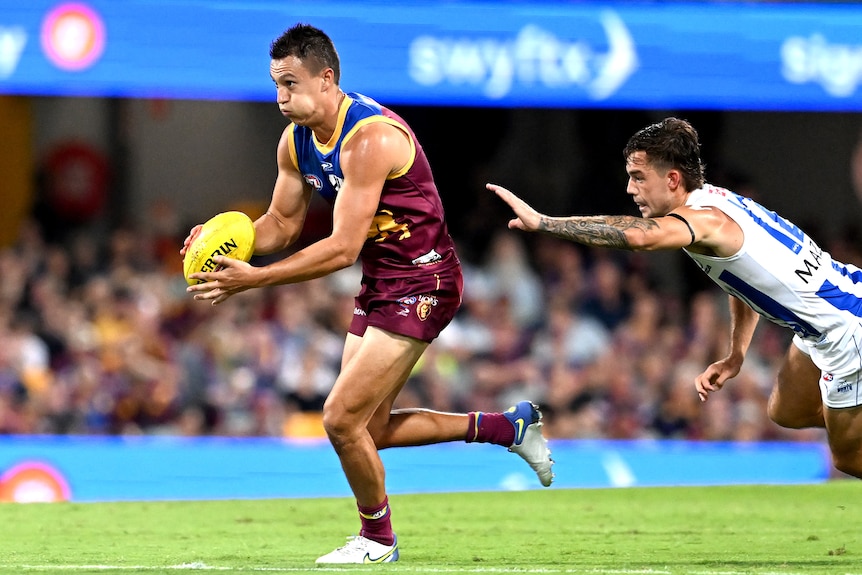 An AFL player puffs his cheeks out as he runs, readying himself to kick the ball while a defender dives at him from behind. 
